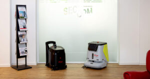 Zwei autonome Reinigungsroboter im Flur eines modernen Bürogebäudes vor einer Glaswand mit dem Firmenlogo von SEC-COM
