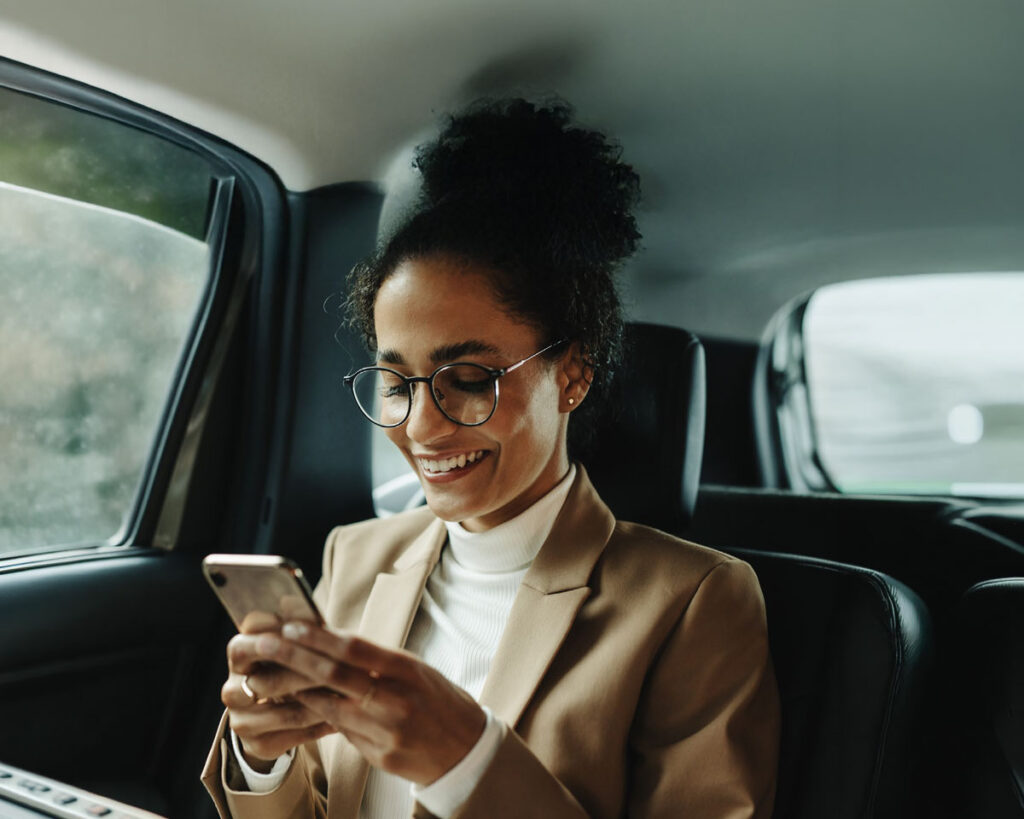 Lächelnde Geschäftsfrau mit Brille sitzt auf der Rückbank eines Autos und nutzt ihr Smartphone. Sie trägt einen beigen Blazer und einen weißen Rollkragenpullover.