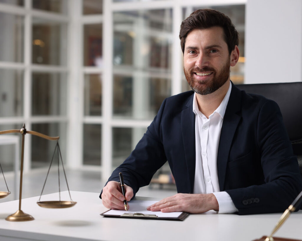 Ein lächelnder Anwalt sitzt in einer modernen Kanzlei an einem Schreibtisch und unterschreibt ein Dokument. Neben ihm steht eine Waage als Symbol der Justiz.