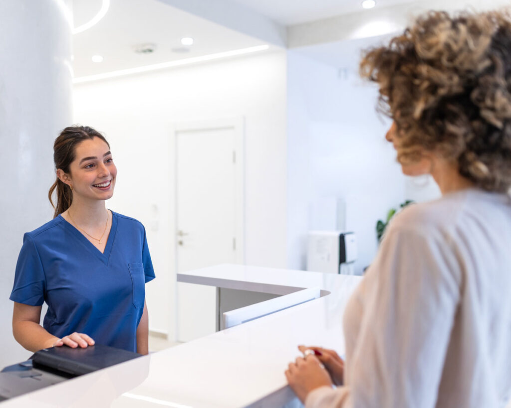 Eine freundliche medizinische Fachangestellte in blauer Arbeitskleidung begrüßt eine Patientin an der Rezeption einer modernen Arztpraxis.