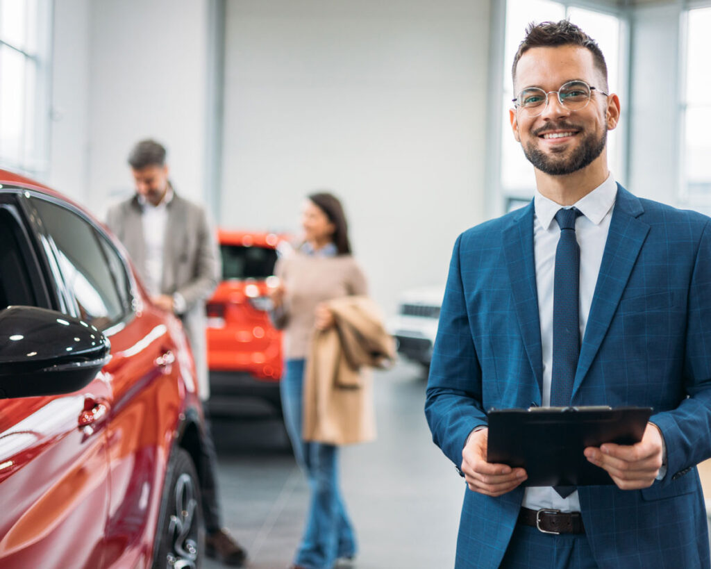Ein freundlich lächelnder Verkäufer im Autohaus steht im Vordergrund mit einem Tablet in der Hand. Neben ihm ist ein rotes Auto zu sehen, während im Hintergrund ein Paar weitere Fahrzeuge betrachtet.